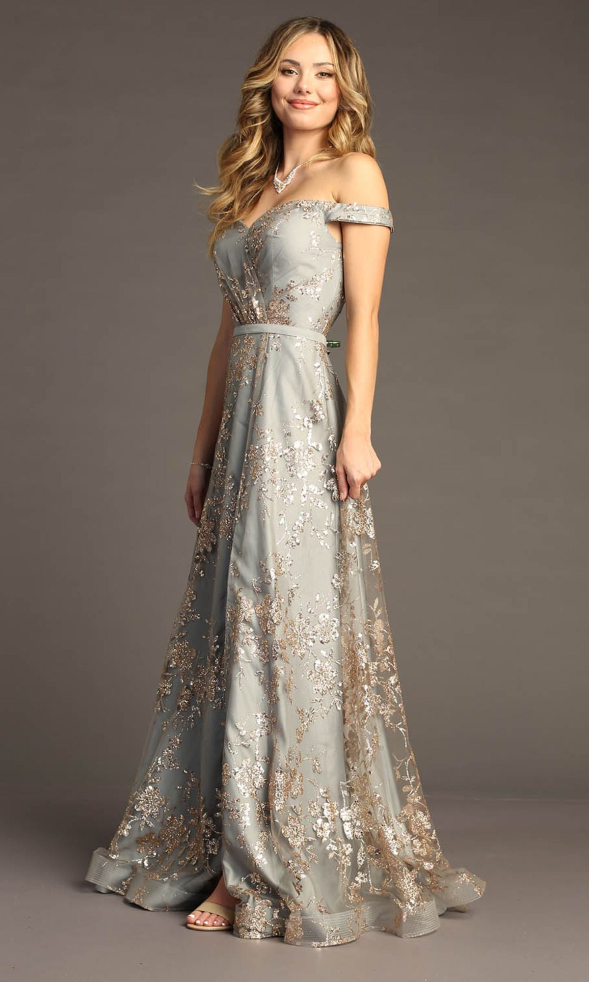 A woman poses elegantly against a neutral background, wearing the Glitter-Print Long Gray Prom Dress: Chicas C6533, which features an off-the-shoulder neckline and sparkling gold floral accents.
